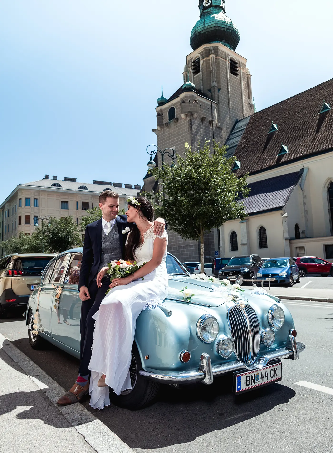 Brautpaar sitzt auf einem Oldtimer vor einer Kirche in stilvoller Hochzeitsfotografie