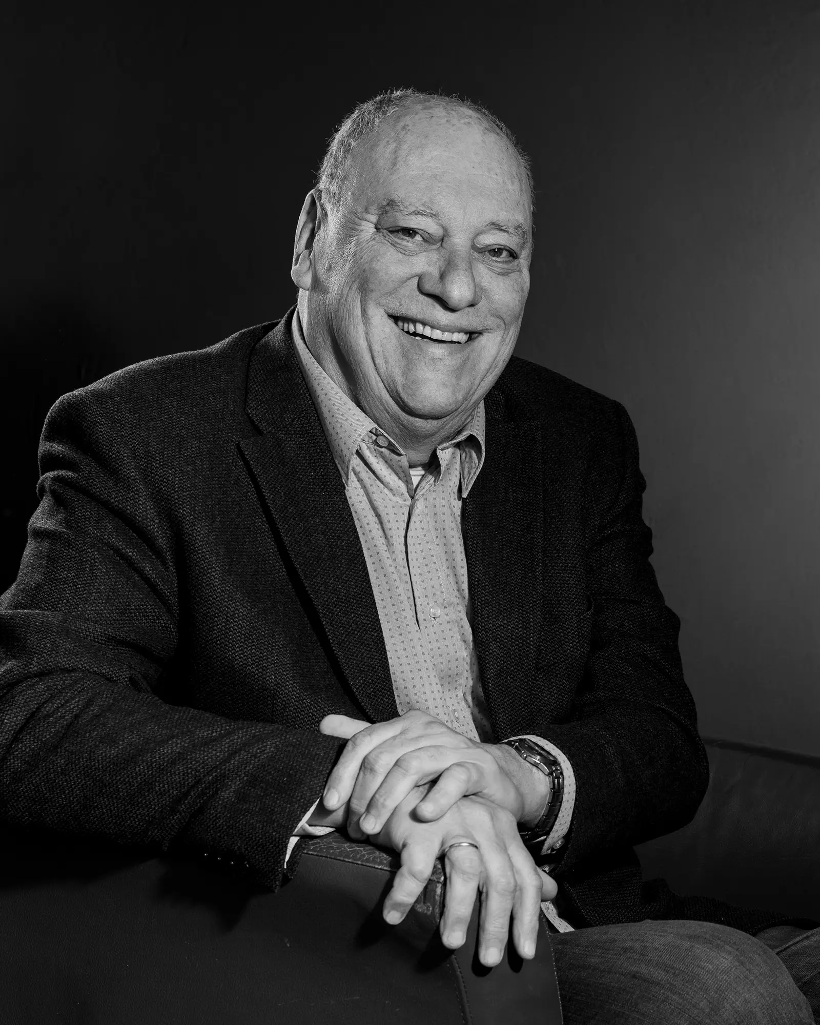 Schwarzweiß Portrait eines lachenden Mannes auf einem Ledersessel mit direktem Blick in die Kamera